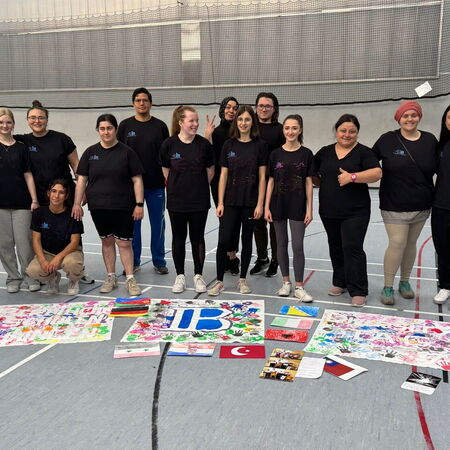 Eine Gruppe von Menschen in schwarzen T-Shirts steht in einer Turnhalle hinter bunten Kunstwerken und einer IB-Karte.