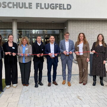 Eine Gruppe von Menschen steht vor der Grundschule Flugfeld und hält ein rotes Band zur Eröffnung oder Feier.