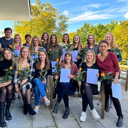 Eine Gruppe von Personen posiert mit Blumen und Zertifikaten für ein Abschlussfoto auf einer Terrasse.