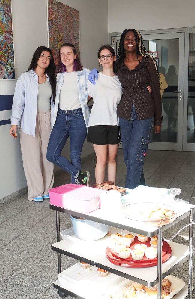 Vier Schüler stehen lächelnd vor einem Tisch mit gebackenem Gebäck in einer Schulumgebung.