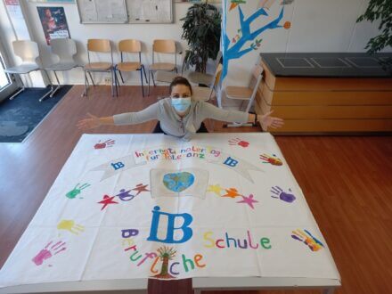 Eine Person mit Maske präsentiert ein buntes Poster mit Handabdrücken und der Aufschrift "IB Schule" in einem hellen Raum.