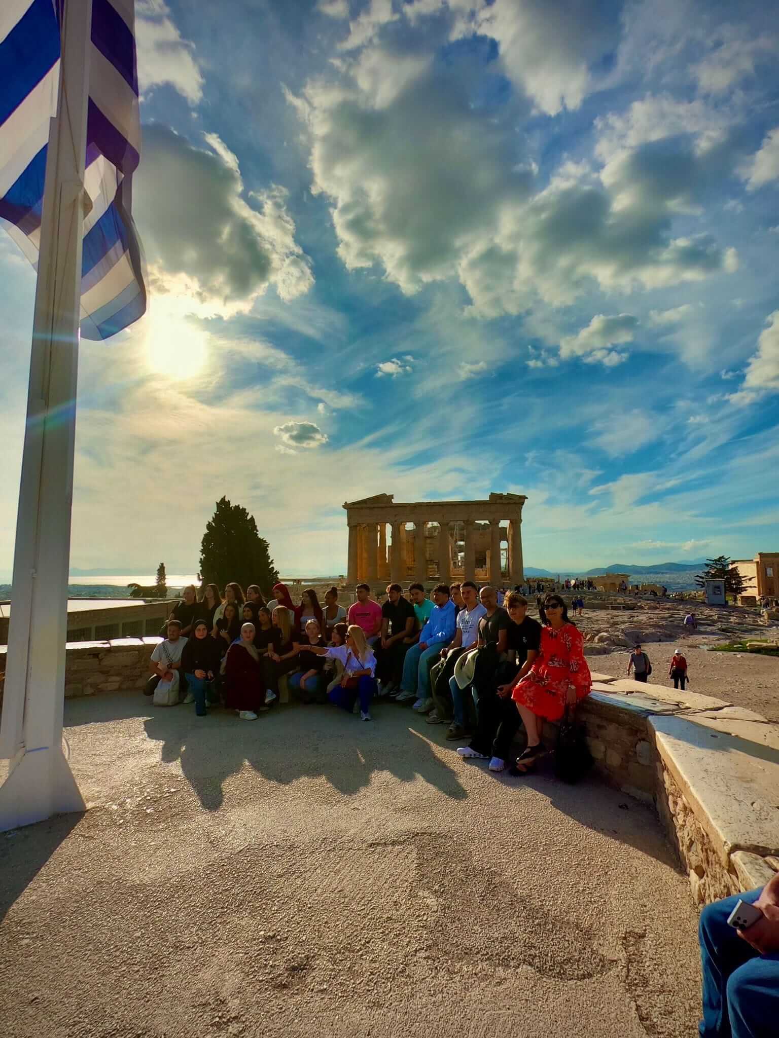 Eine Gruppe von Menschen sitzt neben einer griechischen Flagge mit einem antiken Tempel im Hintergrund bei Sonnenuntergang.